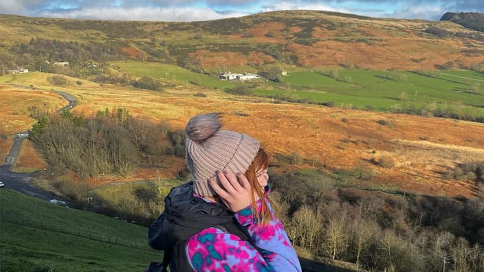 Alex is standing sideways on a mountain wearing a wooly hat. She is pulling her hat down with her hands and there is blue sky and hills in the background.